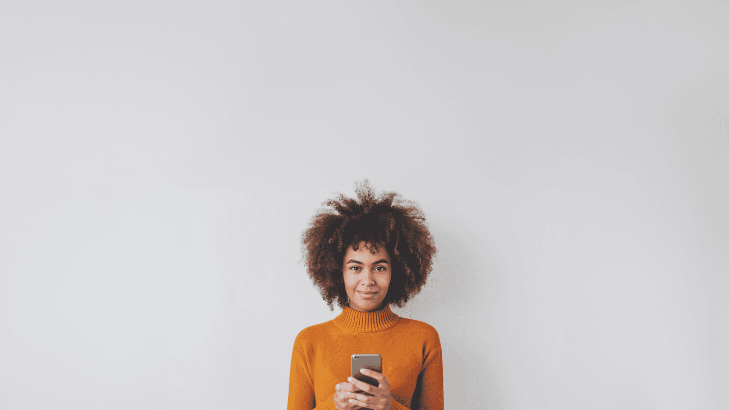 Woman in an orange sweater holding a smartphone against a white wall